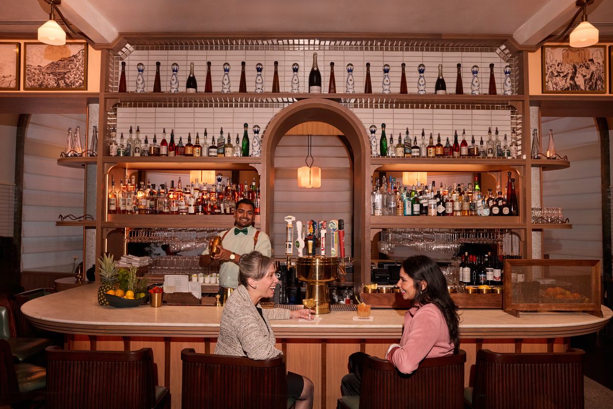 two women sitting at the bar while the bartender makes a drink