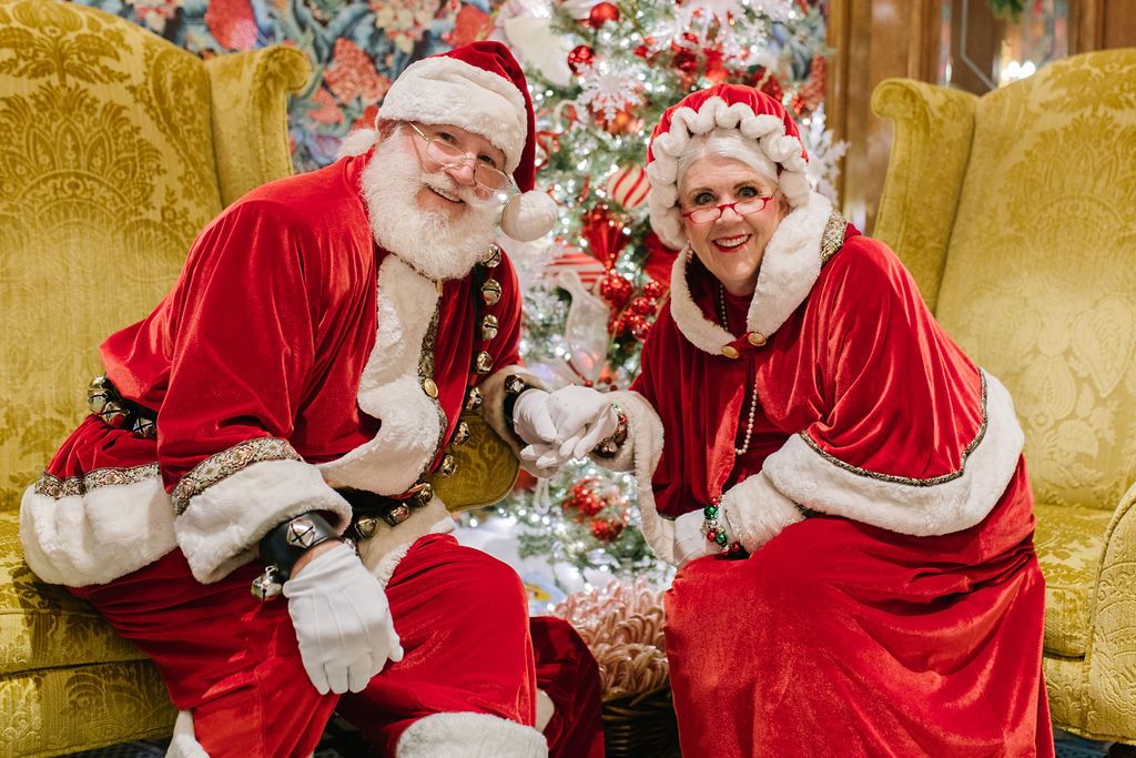 Santa and Mrs. Clause holding hands while sitting in chairs in front of a Christmas tree